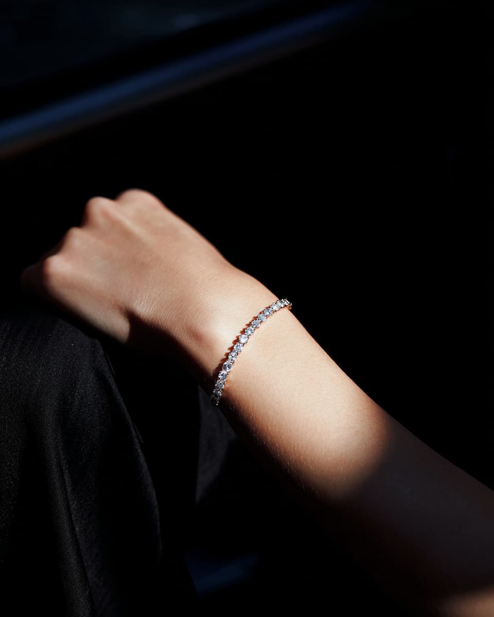 A delicate silver-toned tennis bracelet with small clear stones worn on a person's wrist, shown in dramatic side lighting against a dark background.