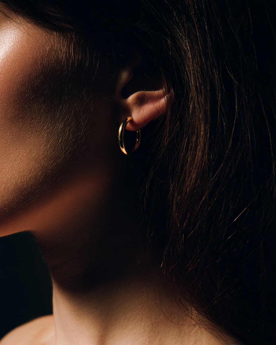A close-up side profile of a woman wearing a small gold hoop earring, with dark hair and warm dramatic lighting against a dark background.