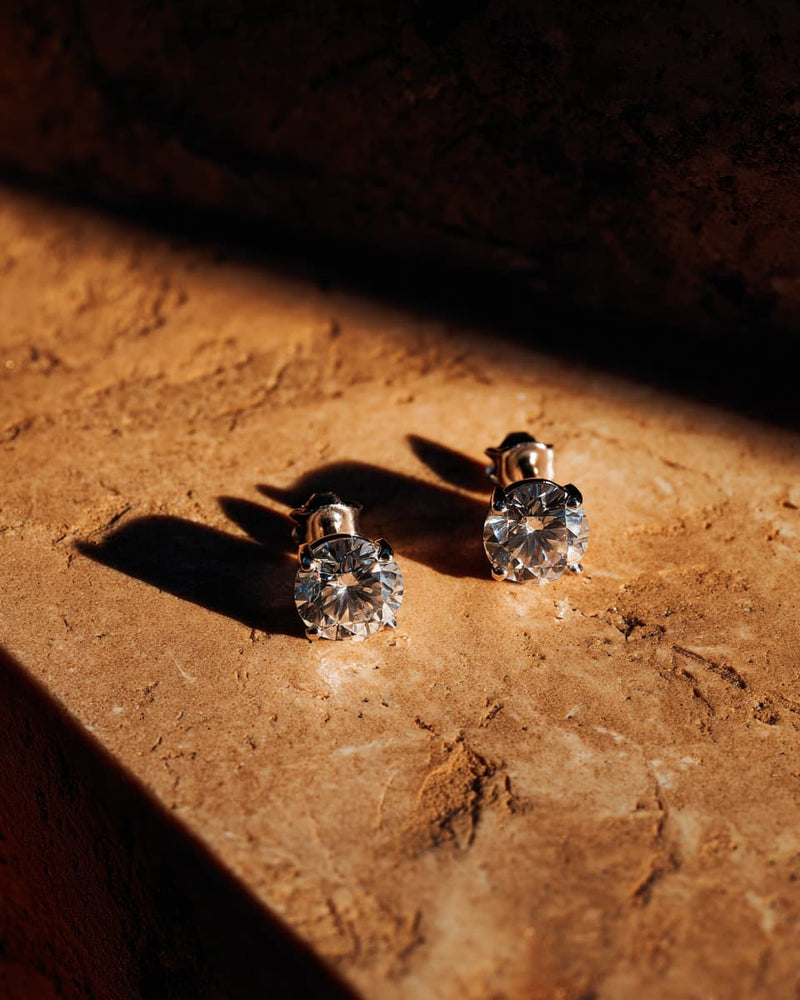 A pair of stud earrings with clear faceted gemstones in gold settings, resting on a textured stone surface under dramatic warm sunlight and shadows.