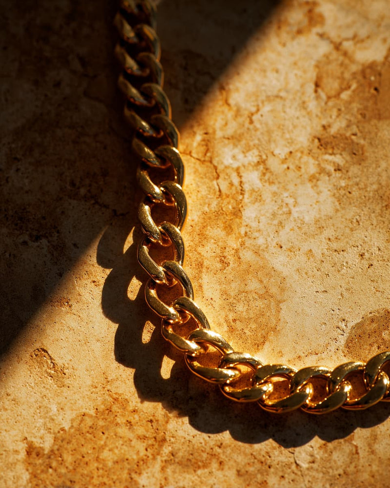 A close-up of a thick gold Cuban link chain draped diagonally across a textured beige stone surface under dramatic warm sunlight and shadows.