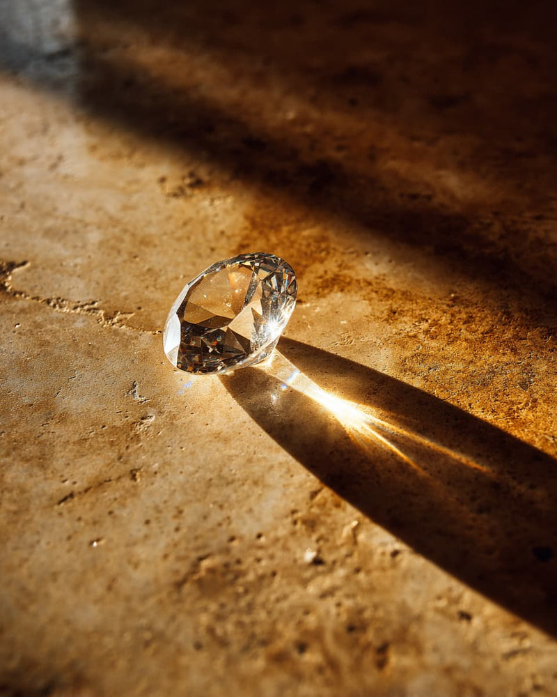 A clear faceted gemstone or crystal resting on a textured warm brown surface, with dramatic sunlight casting a sharp shadow and bright light through the stone.