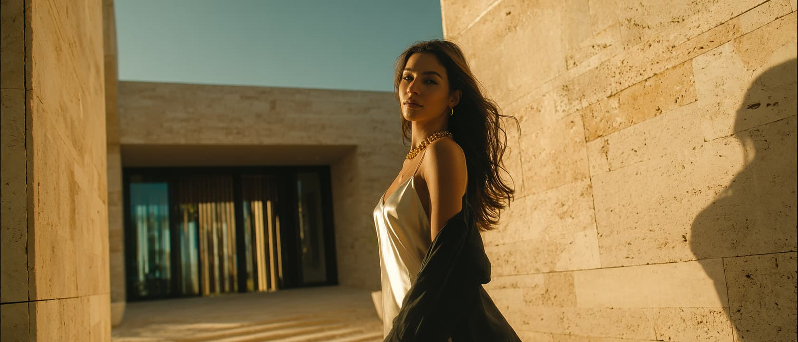 A woman stands outdoors against a sunlit beige stone wall, wearing a metallic silver strapless dress and a dark draped shawl. The scene is warmly lit with strong shadows, and a modern building entrance is visible in the background.
